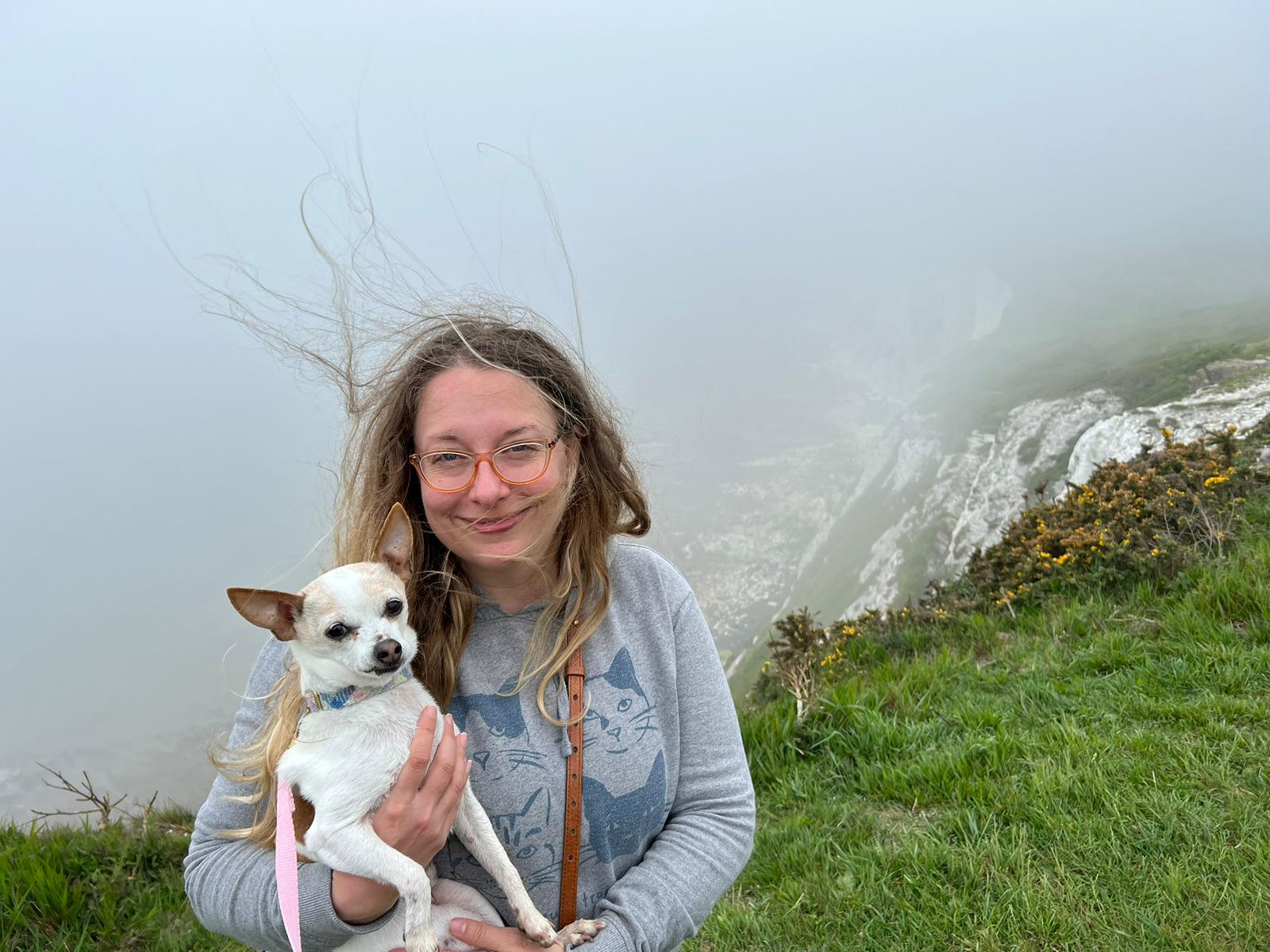 Stacey Kigar smiling outdoors on a misty coastal hilltop, holding her small dog